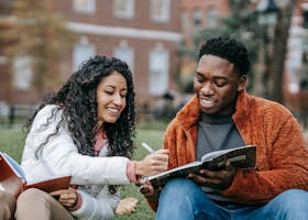 Happy college students studying together on a campus lawn, sharing ideas and writing notes.