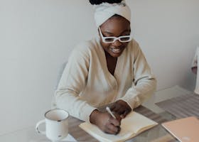 African American woman sitting indoors writing in a journal at a desk with a laptop and coffee mug.
